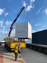 Grúa levantando un contenedor blanco sobre un camión amarillo en un puerto.